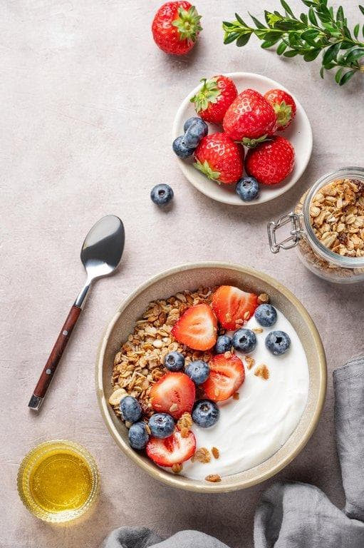 Müsli-Bowl mit Joghurt, frischen Erdbeeren und Blaubeeren, serviert mit Honig und Knuspermüsli in der Draufsicht.