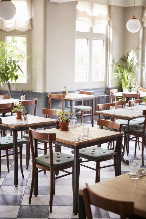 Helles Café mit Holztischen, grünen Polsterstühlen und Schachbrettboden in lichtdurchflutetem Ambiente.