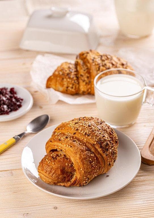 Frisches Laugen-Croissant mit Saaten auf einem Teller, serviert mit einem Glas Milch und Beerenkonfitüre.