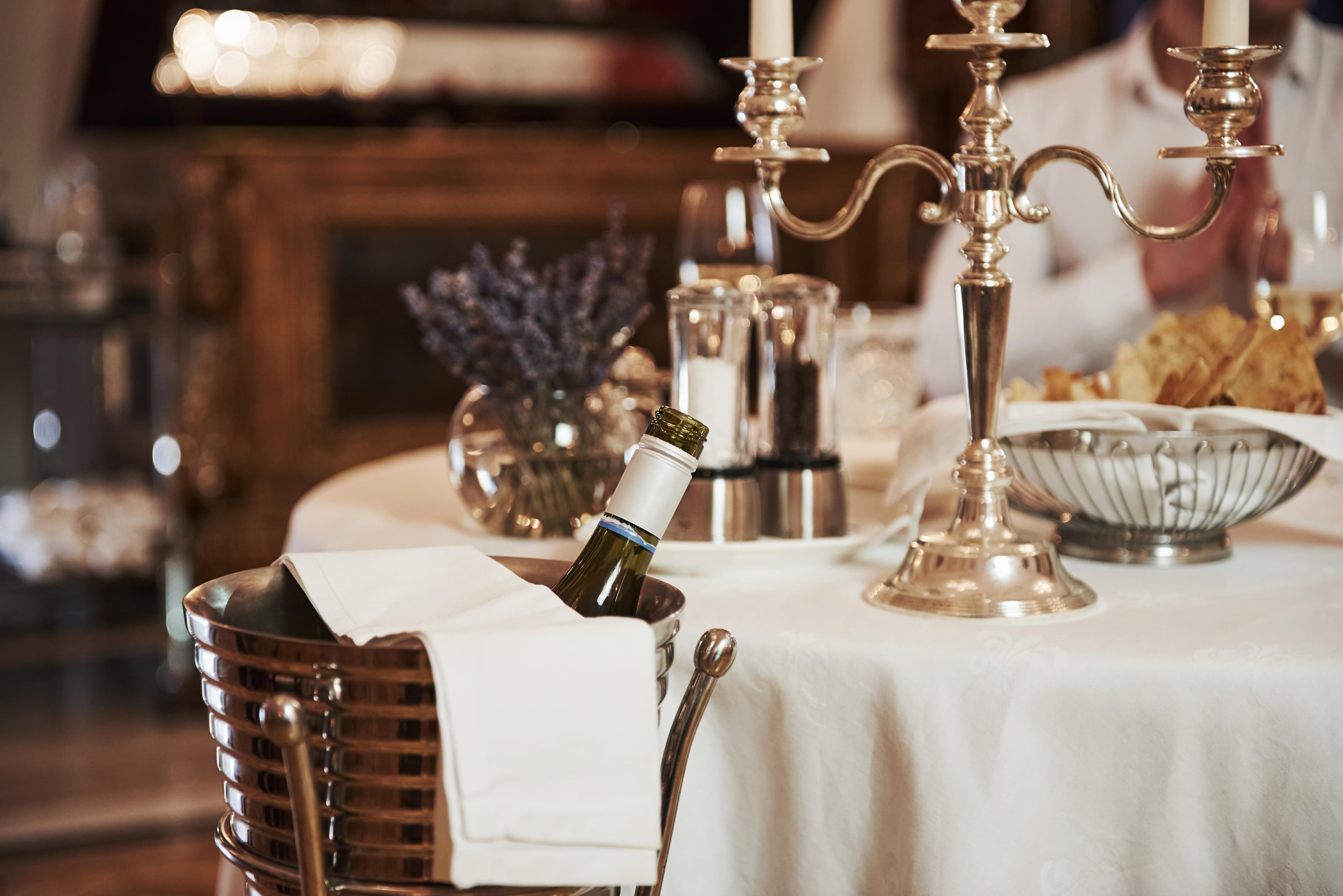 Ein Weinkühler mit einer Flasche Weißwein neben einem elegant gedeckten Tisch mit Kerzenleuchter in einem Restaurant.